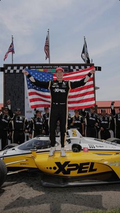 a man standing on top of a yellow race car in front of an american flag