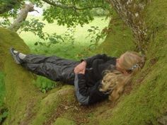 a woman laying on the ground next to a tree and moss covered hill with trees