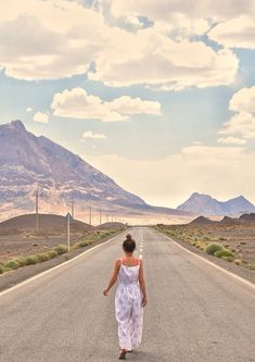 a woman is walking down the middle of an empty road with mountains in the background