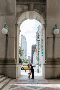 a man and woman standing under an arch in the middle of a city with tall buildings