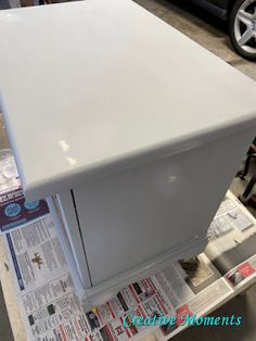 a white refrigerator sitting on top of a table next to a black car and newspaper