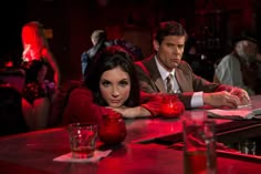 a man and woman sitting at a bar in red latex outfits, with drinks on the table