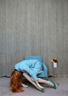 a woman laying on the floor next to a blue chair with her head down and legs bent over