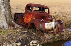 an old rusted truck sitting next to a tree in the middle of a field