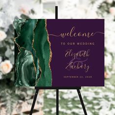 a purple and green wedding welcome sign with gold foil lettering on it, in front of a floral background