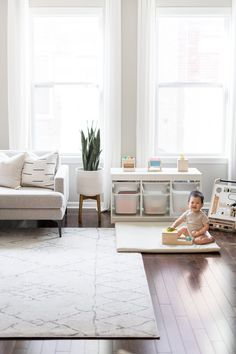 a baby sitting on the floor in front of a living room with white furniture and large windows