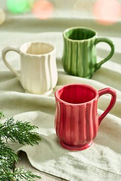 three different colored mugs sitting on top of a white table cloth next to a green plant