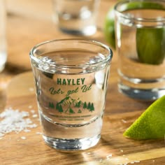 two shot glasses sitting on top of a wooden table next to salt and limes