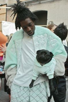 a man with dreadlocks holding a small dog in his arms while walking down the street