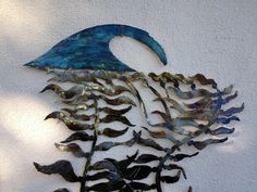 a metal sculpture on the side of a white wall with blue and green leaves hanging from it's sides