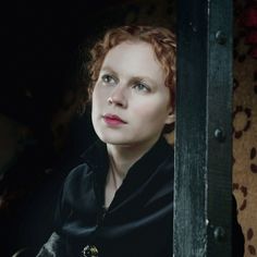 a woman with red hair and blue eyes looking out from behind a metal door in a dark room