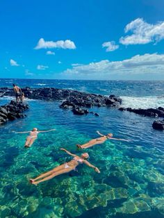 three people are swimming in the ocean with rocks and clear blue water, while one person is standing on his back