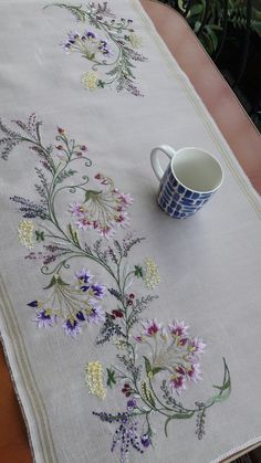 an embroidered table runner with flowers on it and a coffee cup next to it sitting on a wooden table