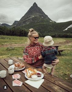 a woman holding a child while sitting at a picnic table in front of a mountain