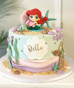 there is a little mermaid sitting on top of a cake with shells and seaweed