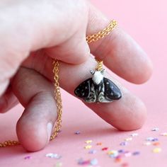 a hand holding a tiny black and white moth pendant on a gold chain with confetti sprinkles around it