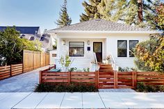 a small white house with wooden fence and trees in the front yard on a sunny day