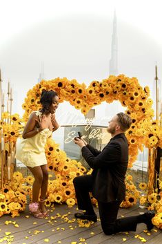 a man kneeling down next to a woman in front of a yellow flowered arch