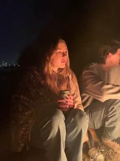 two people sitting next to each other in front of a campfire with the city lights behind them