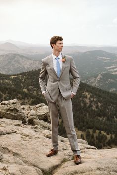 a man in a gray suit standing on top of a rocky hill with mountains in the background