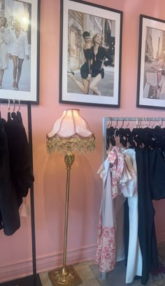 a pink room with pictures on the wall and clothes hanging in racks next to a lamp