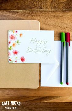 a note card and two pens sitting on top of a wooden table next to an envelope