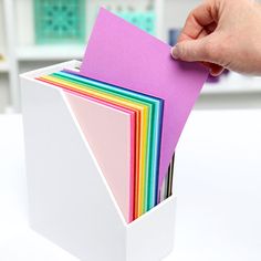 a hand holding a stack of colored papers in a white box on top of a table