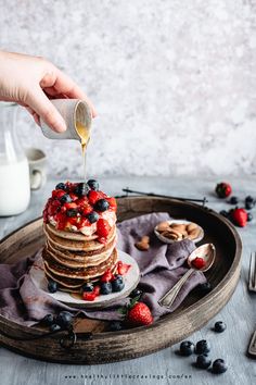 a stack of pancakes with berries and nuts being drizzled with maple syrup