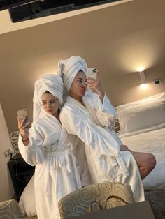 two women in white robes are taking a selfie with their cell phones while sitting on a bed