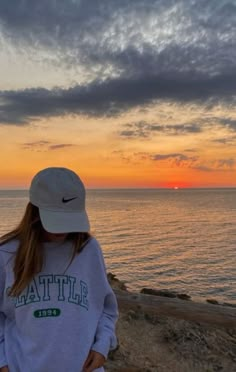 a woman standing on top of a cliff next to the ocean at sunset with her hands in her pockets