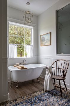 a white bath tub sitting under a window next to a wooden chair and rug on top of a hard wood floor