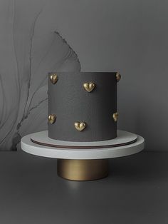 a grey and gold cake with hearts on it sitting on a white stand next to a gray wall