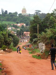 two people walking down a dirt road in front of some houses and trees with a church on the hill behind them