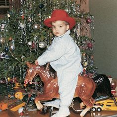 a little boy in a cowboy hat riding a toy horse next to a christmas tree