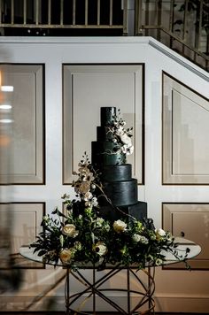 a black wedding cake with white flowers and greenery sits on a table in front of a staircase