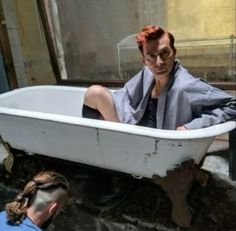 a woman sitting in a bathtub with her feet up on the ground while another man looks at her