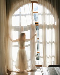 a woman standing in front of a window with her arms out and hands on the curtains