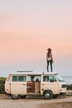 a woman standing on top of an old vw van with the ocean in the background