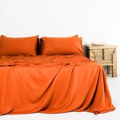 a bed with orange sheets and pillows next to a night stand in a white room