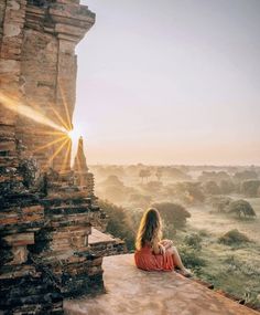 a woman sitting on top of a stone wall next to a building with the sun shining down