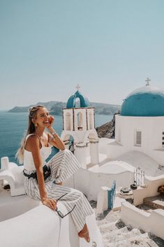 a woman sitting on top of a white building next to the ocean with blue domes