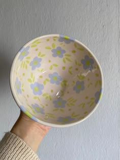 a hand holding a bowl with blue and green flowers on the bottom, in front of a white wall