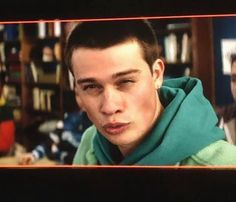 a young man in a green hoodie stares at the camera while standing next to a bookshelf