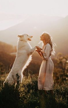 a woman standing next to a white dog on top of a lush green field with mountains in the background