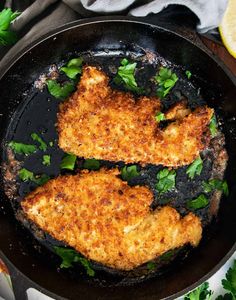 two pieces of chicken cooking in a skillet with parsley and lemon wedges