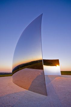 a large metal object sitting in the middle of a field at sunset or dawn with its reflection on it's surface