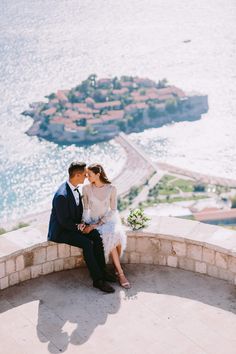 a man and woman are sitting on a wall near the water with an island in the background