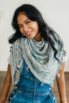 a woman wearing overalls and a knitted shawl smiles at the camera with her hands in her pockets