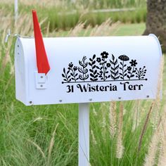 a white mailbox with black and red flowers on it in front of some tall grass