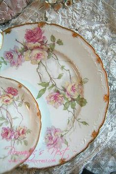 two plates with pink flowers on them sitting on a lace covered tablecloth next to silverware